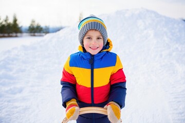 Joyful child in a bright jacket holds a sled on a snowy hill for World Snow Day