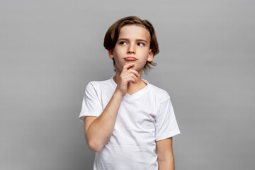 Pensive young boy looking away in studio with thoughtful expression on face. Child model wearing white t-shirt posing against gray background indoors