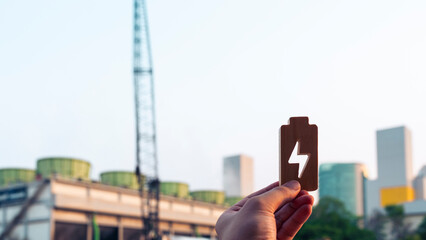 Hand holding wooden battery icon with energy symbol in front of power plant and modern buildings. Concept for renewable energy, sustainable power, eco technology, and electricity industry.