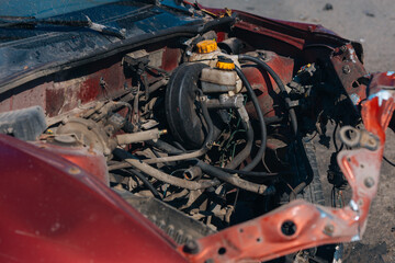 A serious road accident: a wrecked red car with a severely damaged front end and a flat tire sits on the pavement. The car shows signs of a head-on collision, including a destroyed engine and hood.