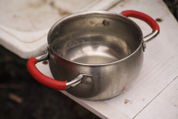A dirty small stainless steel pot with red rubber handles sits on a dirty white surface next to a sink.