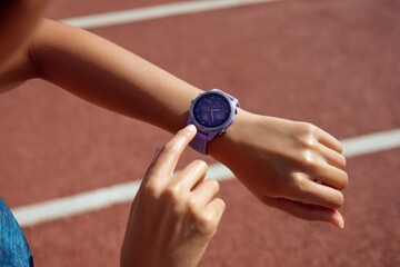 Close-up of a runner tapping a lavender sports smartwatch while standing on the track. Bright sunlight enhances the sleek design and athletic mood of the training moment.