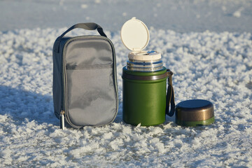 Green food thermos with cup, cover and spoon in winter on snow