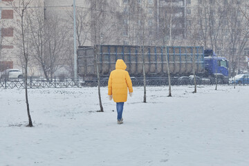 A woman in a yellow jacket walks through white snow with a truck in the background in winter.