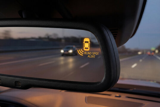 Car rearview mirror displaying digital blind spot alert warning icon during evening highway drive