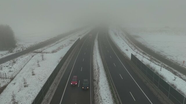 Moving cars on a winter expressway in dense fog with snowy roadsides and noise barriers captured from a drone