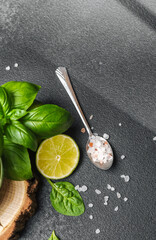 Fresh green basil leaves, lime slice, and salt crystals on spoon scattered on dark slate background