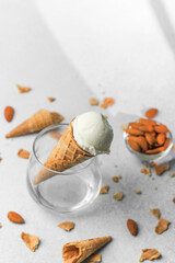 Vanilla ice cream in waffle cone placed in clear glass with almonds and broken waffle pieces on grey background