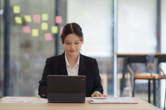 An Asian businesswoman smiling and working on a digital tablet at an office desk.
