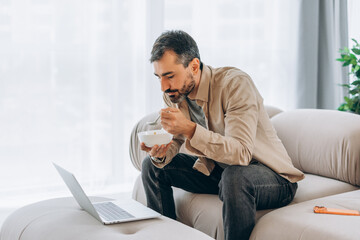 Middle aged bearded man eating breakfast while working on laptop at home
