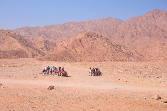 Tourists riding camels in desert landscape with mountains and clear blue sky in background