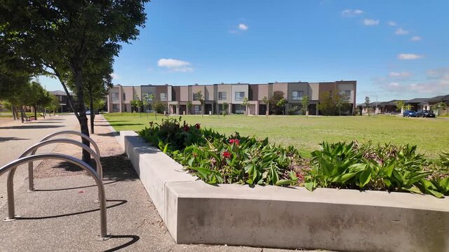 A landscaped public space in Tarneit, Melbourne, Australia, featuring modern townhouses overlooking a large green lawn, young street trees. Concept of suburban community design, residential planning