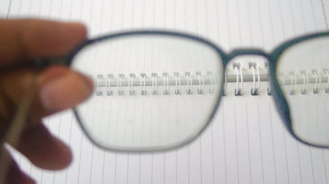 Close up of a hand holding eyeglasses in front of an eye chart on a notebook. The glasses are focusing on different lines of the chart, illustrating vision correction and eye health concept.