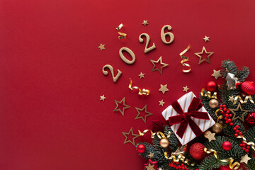 Gift box and 2026 numbers for New Year celebration background in red color with fir branches, red and gold balls, berries and festive baubles, top view