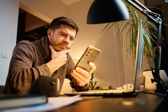 Man sitting at desk using smartphone while working in a home office with laptop and documents. Tired freelancer procrastinating at workplace during remote work. Unproductive man using phone