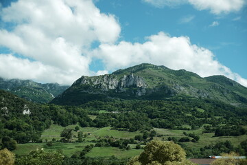 Abstract landscape in northern Spain, with green meadows and trees in the foreground and partially vegetated mountains under a blue sky with clouds. Rural peaceful landscape