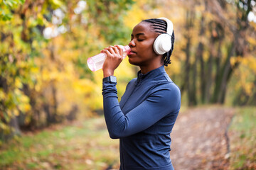 Black woman in athletic wear standing in park in autumn, drinking water after jogging.
