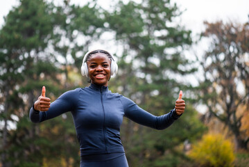 Black woman who is ready for jogging stands in park in winter, looking relaxed and happy, showing thumbs up.