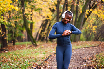 A runner in dark athletic wear moves confidently down a wooded path, framed by vibrant fall foliage and soft natural light. She is looking at smartwatch.