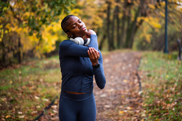 Young black woman is exercising in park in autumn. She is stretching her body before jogging.
