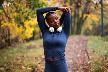 Young black woman is exercising in park in autumn. She is stretching her body before jogging.