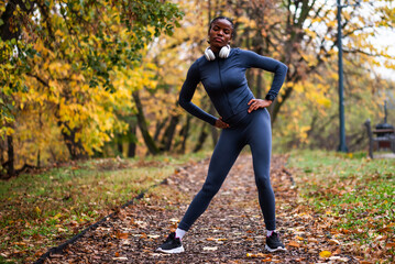 Young black woman is exercising in park in autumn. She is stretching her body before jogging.