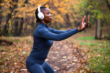 Young black woman is exercising Tai chi exercise in park in autumn. She is wearing wireless headphones.