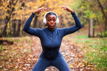 Young black woman is exercising Tai chi exercise in park in autumn. She is wearing wireless headphones.
