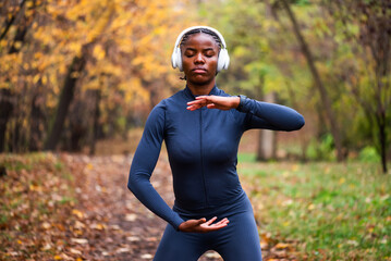 Young black woman is exercising Tai chi exercise in park in autumn. She is wearing wireless headphones.