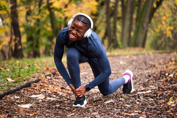 Young black woman is jogging in park in autumn and got injured. She has pain in her ankle.