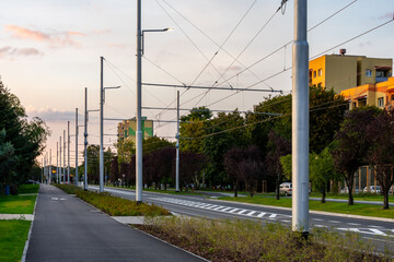 Fototapeta premium Urban Tram Wires And Street Poles Along Green City Road At Sunset In Modern Transportation Infrastructure