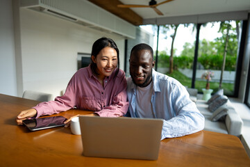 Happy black and Asian couple using laptop together, smiling wife and husband shopping online