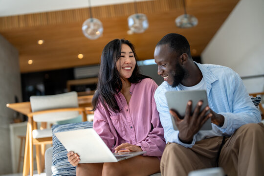 Young happy multiethnic couple having fun while using laptop, digital device at home