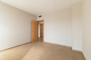 Empty room with beige walls and wooden door. The floor is tiled in light brown. Natural light enters through the open door, creating a spacious feel.