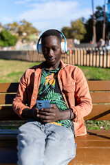 Fototapeta premium Young African man enjoying leisure time on a park bench, listening to music and using smartphone
