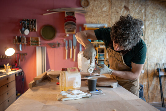 Man carpenter preparing for work on new furniture object, precisely measures required amount of epoxy resin on electronic scales at table, pouring liquid in paper cup from canister, workshop process. - Powered by Adobe