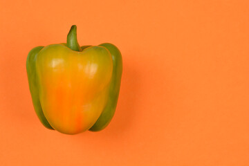 A fresh bell pepper showing a natural yellow-green color transition is positioned on the left side of a solid orange background