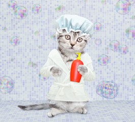 Cute kitten with shower cap on it head wearing bathrobe holding bottle of shampoo and standing on hind legs at bathroom at home
