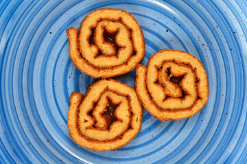 Three slices of spiral sponge cake filled with dark jam are arranged on a blue ceramic plate with a concentric ring pattern