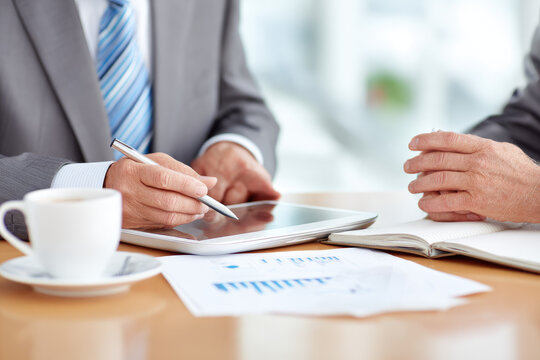 Senior businessman mentoring young employee during meeting, discussing financial data with digital tablet and documents on office desk, professional atmosphere, guidance and support