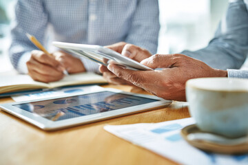 Senior businessman mentoring young employee in modern office using digital tablet and discussing financial report with coffee on wooden desk