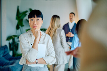 Asian businesswoman thinking diligently, holding tablet and smartphone, listening to colleagues in a corporate office discussion
