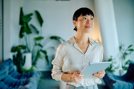 Asian businesswoman holding a modern digital tablet, looking away with a thoughtful expression, planning innovation and bright future