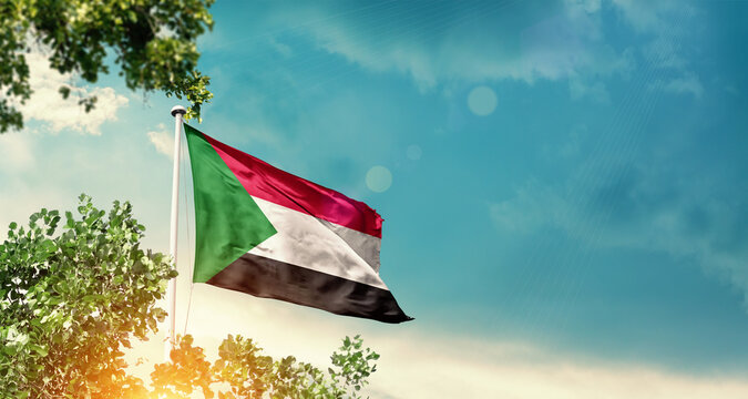 Sudan national flag waving in the sky behind the tree. The flag blowing with beautiful sunlight. Sudan national flag for independence day. - Powered by Adobe