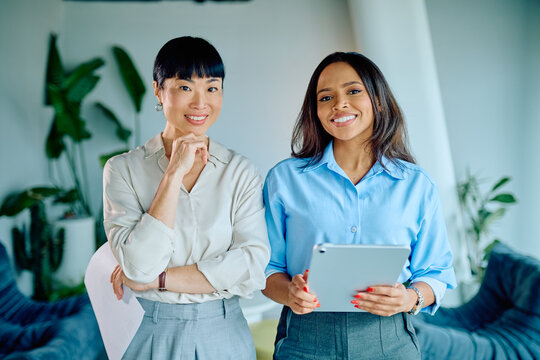Two professional multi-ethnic businesswomen standing together, smiling and holding a digital tablet in a modern office environment - Powered by Adobe