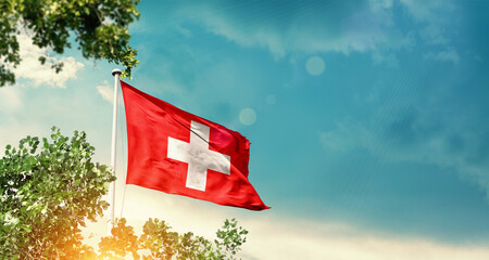 Switzerland national flag waving in the sky behind the tree. The flag blowing with beautiful sunlight. Switzerland national flag for independence day.