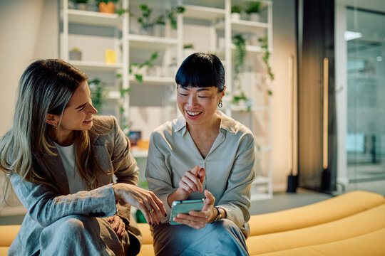 Two smiling businesswomen interacting with a mobile phone, sharing a laugh and content while relaxing in a modern office lounge area