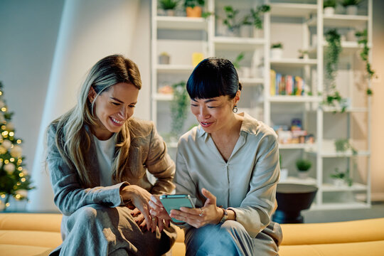 Two diverse female coworkers looking at a smartphone, discussing content and sharing a happy moment in a contemporary office setting