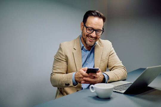 Happy businessman smiling, texting, or browsing on mobile phone, sitting at an office desk with a laptop and coffee