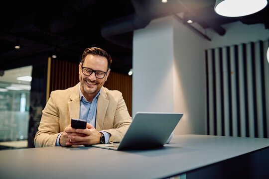 Smiling businessman working remotely, holding mobile phone and looking at screen, with laptop on table in an contemporary workspace
